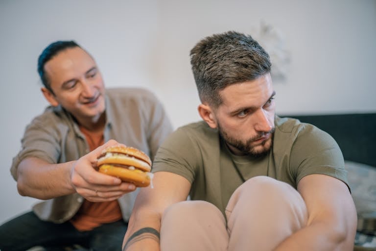 A man humorously offers a burger to his friend who seems uninterested or upset.